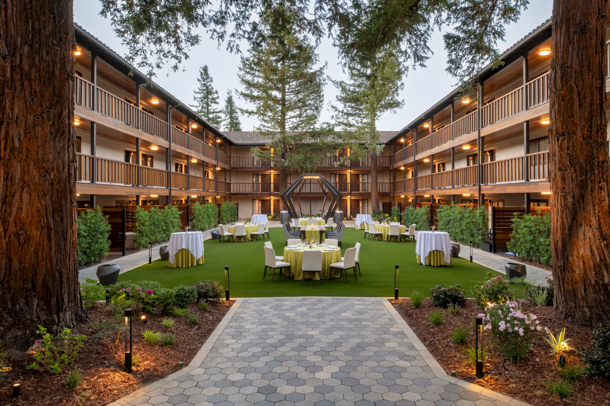 The image shows a courtyard with tables and chairs arranged on grass, surrounded by a three-story building with balconies and tall trees.