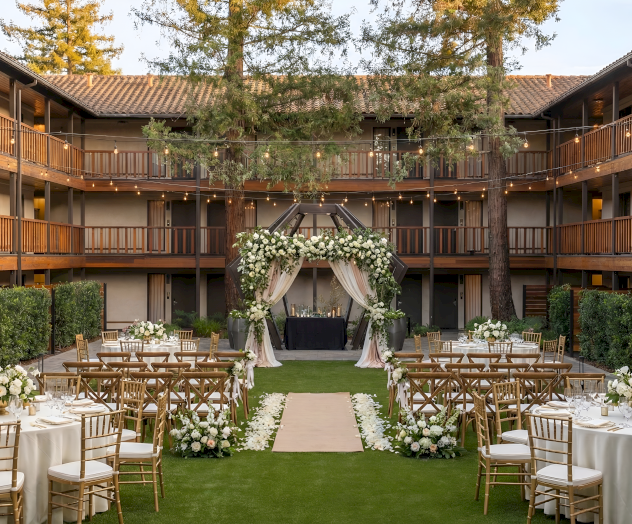A wedding setup on a courtyard lawn with round tables, wooden chairs, floral arrangements, and a draped archway under trees.