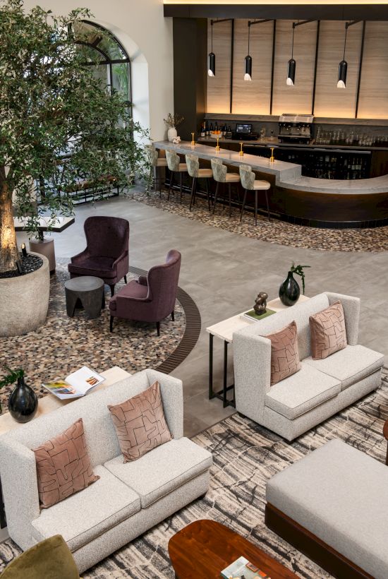 A modern hotel lobby with a curved bar, warm lighting, tall bar stools, beige sofas, neutral cushions, potted trees, and a cozy seating area.