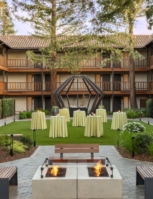 An upscale hotel courtyard with a square fire pit, modern seating, tall trees, and round cocktail tables set for guests under a wooden-lattice building.