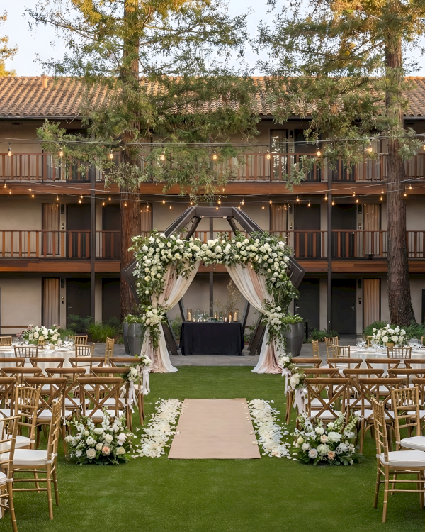 Outdoor wedding setup in a courtyard with a floral arch, wooden chairs, round tables, and elegant centerpieces on green lawn. Ends right.