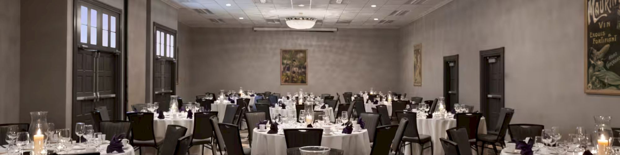 A banquet hall set for an event with round tables, white tablecloths, glassware, and candles, surrounded by patterned chairs.