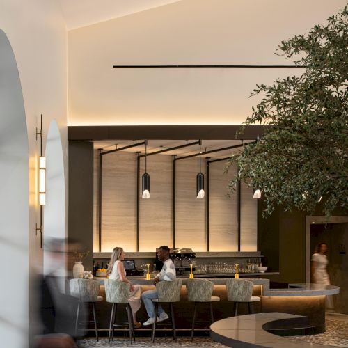 A modern hotel lobby with a long bar, pendant lights, and people seated at a curved counter, while a tree and round table accent the space.