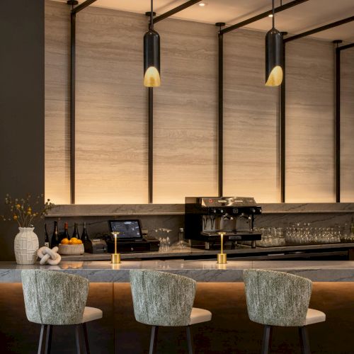 A chic bar interior with a dark wood counter, three upholstered stools, pendant lights, and a sleek coffee/ beverage setup&mdash;stylish and inviting.