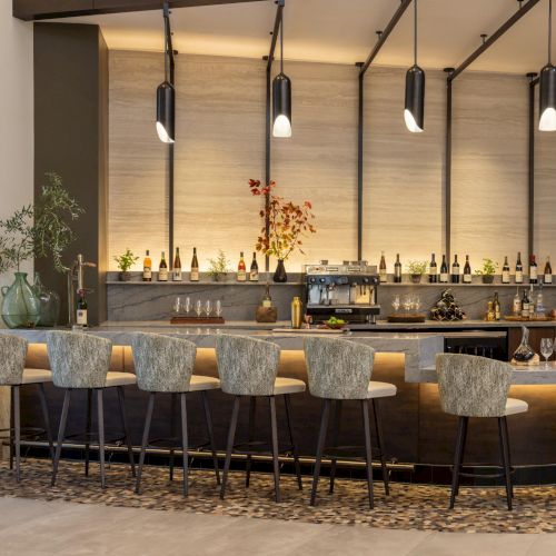 A modern bar with a long wooden counter, glassware, pendant lights, and cushioned stools, set against a sleek, backlit shelf and patterned floor, design-forward.
