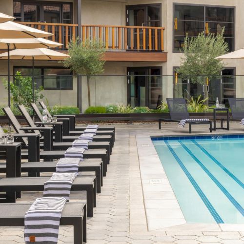 A hotel pool area with many lounge chairs, umbrellas, and a clear blue pool by a courtyard building.