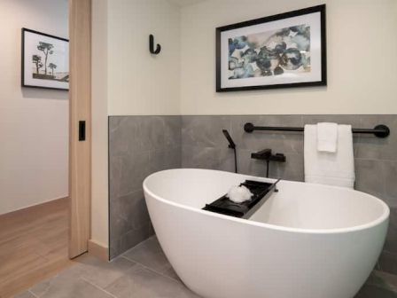 A modern bathroom with a freestanding white bathtub, wall-mounted faucet, towel bar, and framed artwork on the walls, plus neutral tiles.