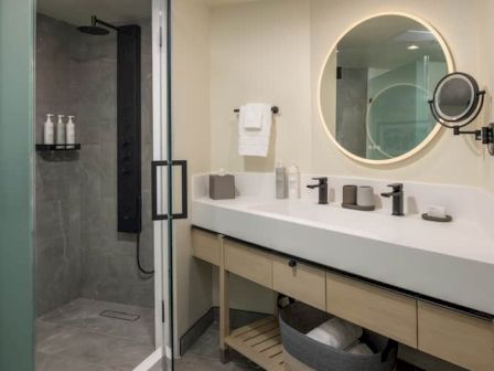 A modern hotel bathroom with a glass shower, round backlit mirror, double sink, and tidy toiletries, plus a small basket under the counter.