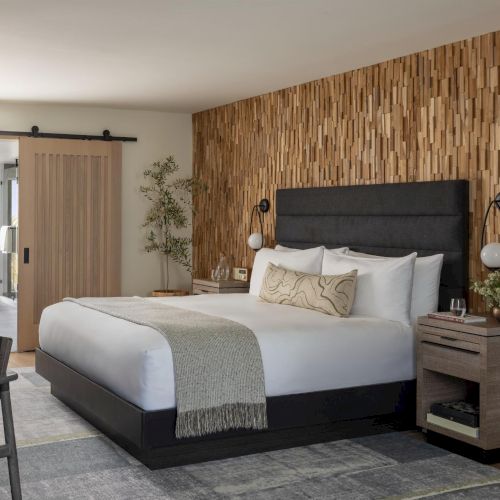 A modern hotel room with a stone-accent wall, king bed, nightstands, and a view into a bathroom and balcony. Top it at 140 characters, ending the sentence.