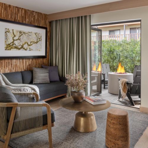 A modern living room with a gray sofa, wooden accents, a round coffee table, and a sliding glass door opening to a patio with a fire feature outside.
