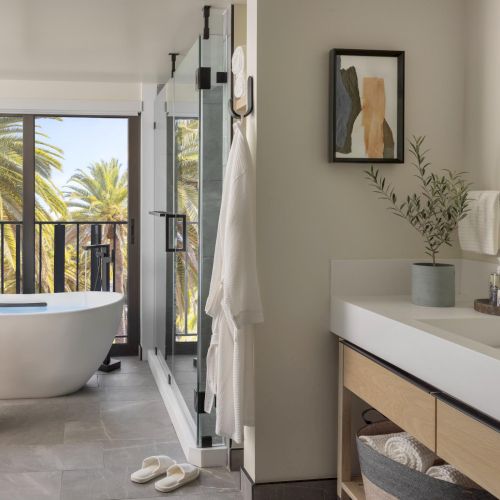 A modern bathroom with a freestanding tub by a glass door, a separate shower, a wooden stool, and a double sink vanity with round mirror, airy and bright.