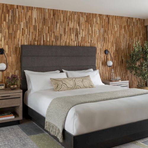A stylish hotel bedroom with a textured wood accent wall, upholstered bed, gray headboard, two nightstands, wall sconces, a TV, and a potted plant by the window.