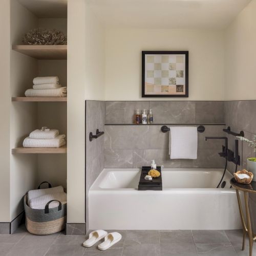 A modern bathroom with a white bathtub, tiled surround, open shelving with towels, a small side table, and a balcony view outside, neat and airy.