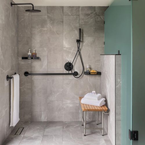 A modern, accessible shower with gray tiled walls and floor, a wall-mounted rainfall head, a handheld wand, a towel bar, a small bench with towels, and a glass door.