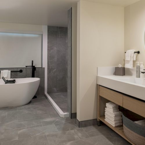 A modern bathroom with a freestanding tub, glass shower, and a double-sink vanity with round mirror, neutral tones, and tidy shelves, end.