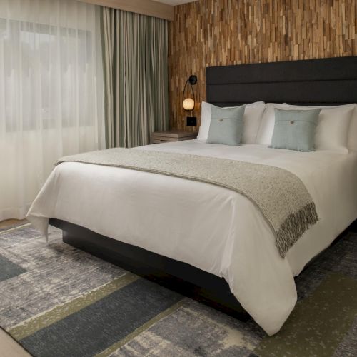 A modern hotel bedroom with a king bed, neutral tones, and pendant lights by two nightstands, plus a patterned rug.