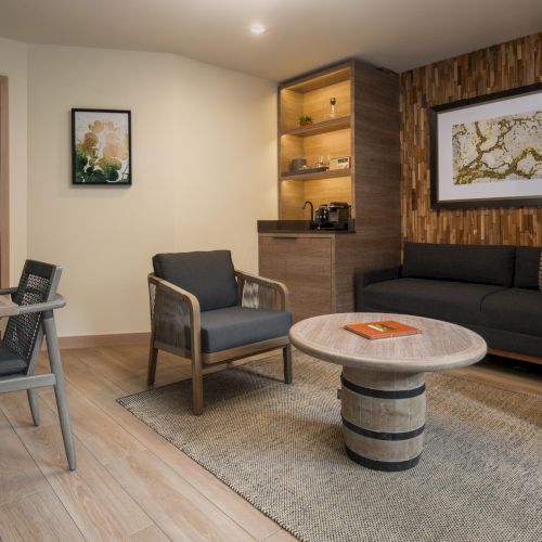 A modern, cozy living room with a gray sofa, wooden armchairs, round stone-top coffee table, a built-in shelf, and warm wood accents.
