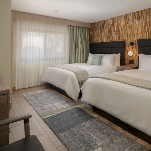 A hotel room with two large beds, modern wooden accent wall, soft lighting, a window with sheer curtains, a dresser, and a patterned rug, tidy and inviting.