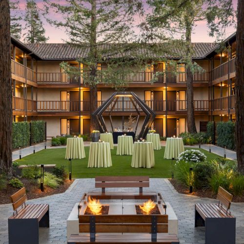 A glass-roofed, plant-filled atrium with a brick patio, lush greenery, and a central seating area flanked by trees.